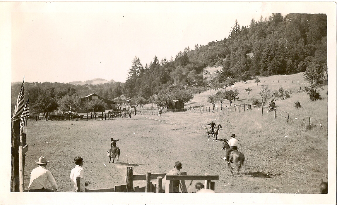 A historical black and white image shows four horseback riders riding in a corral situated within a valley, as spectators watch the riders in the foreground.
